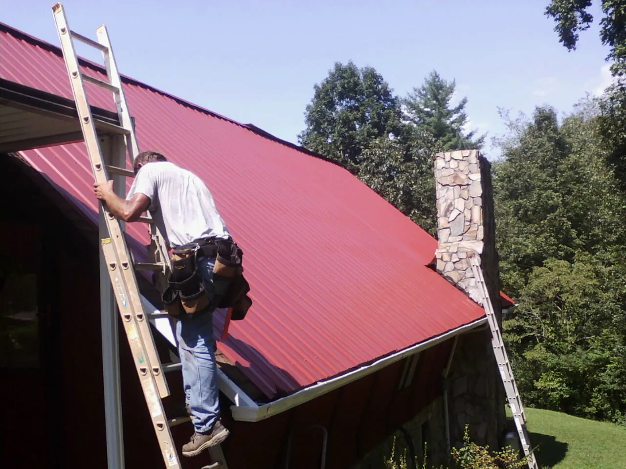 Roofer on a ladder beside a red metal roof and stone chimney — Soffit & Fascia Repair by Roofing Brothers in Hammondsport